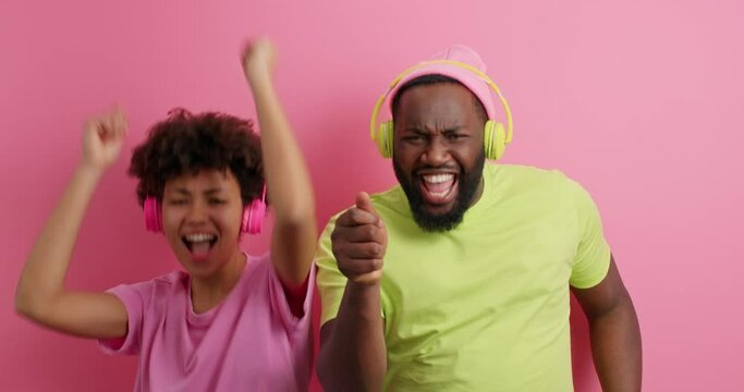 Happy dark skinned couple jump high raise arms feel overjoyed listen music via wirelesss headphones dressed in casual t shirts isolated over pink background. People leisure amusement concept