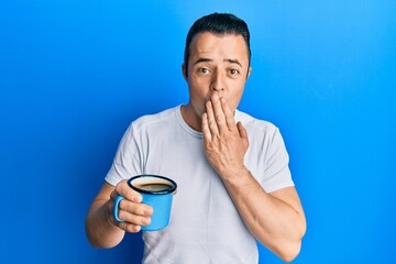 Handsome young man drinking a cup coffee covering mouth with hand, shocked and afraid for mistake. surprised expression