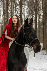 beautiful girl in a white dress and a red cloak with a black horse in the spring forest