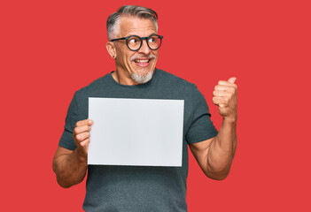 Middle age grey-haired man holding blank empty banner pointing thumb up to the side smiling happy with open mouth