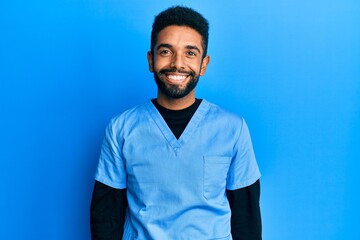 Handsome hispanic man with beard wearing blue male nurse uniform looking positive and happy standing and smiling with a confident smile showing teeth