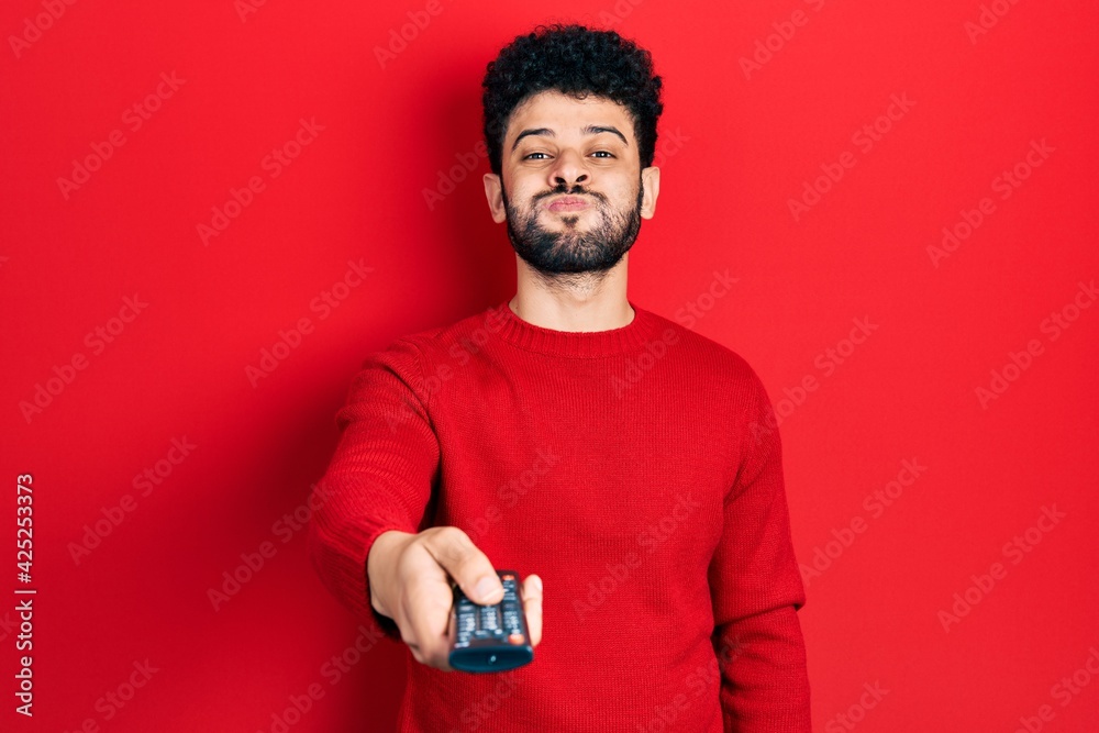 Poster young arab man with beard holding television remote control puffing cheeks with funny face. mouth in