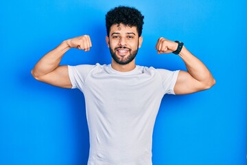 Young arab man with beard wearing casual white t shirt showing arms muscles smiling proud. fitness concept.