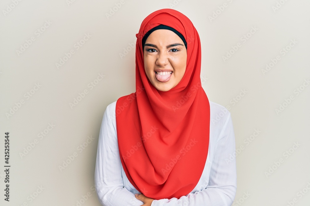 Wall mural young beautiful hispanic girl wearing traditional islamic hijab scarf sticking tongue out happy with