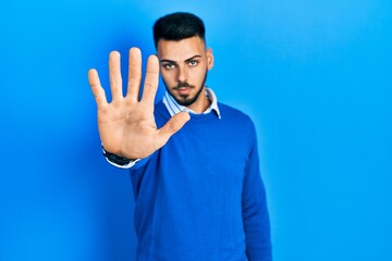 Fototapeta premium Young hispanic man with beard wearing casual blue sweater doing stop sing with palm of the hand. warning expression with negative and serious gesture on the face.
