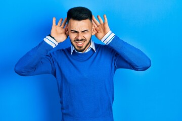 Young hispanic man with beard wearing casual blue sweater trying to hear both hands on ear gesture, curious for gossip. hearing problem, deaf