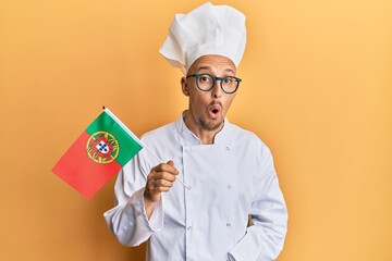Bald man with beard wearing professional cook apron holding portugal flag scared and amazed with open mouth for surprise, disbelief face