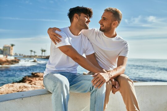 Young gay couple smiling happy sitting on the bench at the beach promenade.