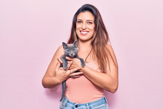 Young beautiful latin woman smilling happy. Standing with smile on face holding adorable cat over isolated pink background
