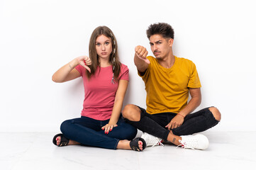 Fototapeta na wymiar Young couple sitting on the floor isolated on white background showing thumb down with negative expression