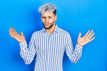 Naklejka premium Young hispanic man with modern dyed hair wearing business striped shirt clueless and confused with open arms, no idea and doubtful face.
