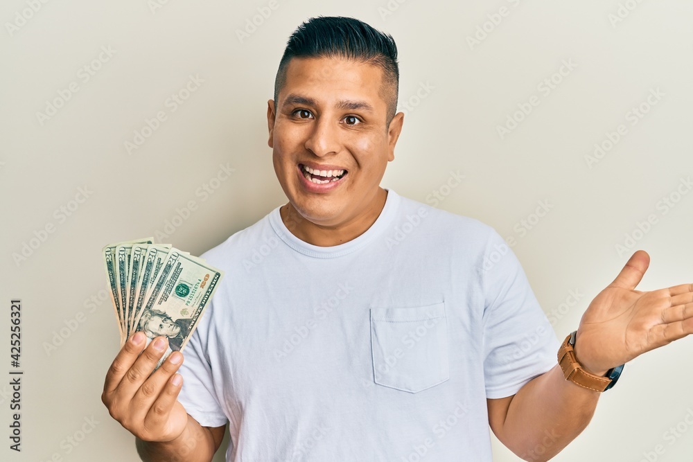 Wall mural young latin man holding 20 dollars banknotes celebrating achievement with happy smile and winner exp