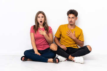 Fototapeta na wymiar Young couple sitting on the floor isolated on white background surprised and shocked while looking right