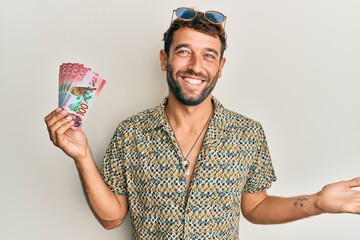 Handsome man with beard holding 100 new zealand dollars banknote celebrating achievement with happy smile and winner expression with raised hand