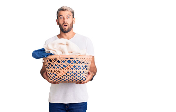 Young handsome blond man holding laundry basket with clothes scared and amazed with open mouth for surprise, disbelief face