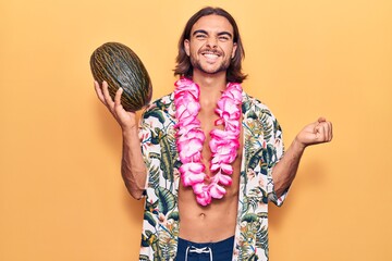 Young handsome man wearing swimwear and hawaiian lei holding melon screaming proud, celebrating...
