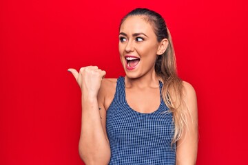 Young beautiful blonde woman wearing sporty t-shirt standing over isolated red background pointing thumb up to the side smiling happy with open mouth