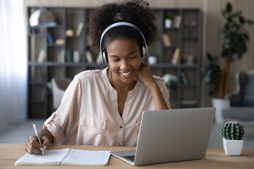Smiling young African American female in headphones write look at laptop screen study online on gadget. Happy millennial biracial female in earphones work distant on computer. Education concept.