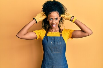 Middle age african american woman wearing professional apron smiling pulling ears with fingers,...