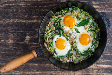 Shakshuka, fried eggs with green wild garlic leaves, onion, pepper and spices in cast iron pan, close up