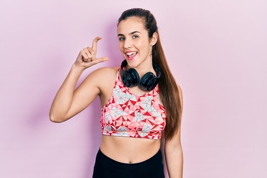 Young brunette teenager wearing gym clothes and using headphones smiling and confident gesturing with hand doing small size sign with fingers looking and the camera. measure concept.