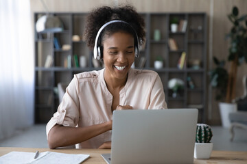 Smiling young African American woman in headphones work on laptop have webcam online digital...