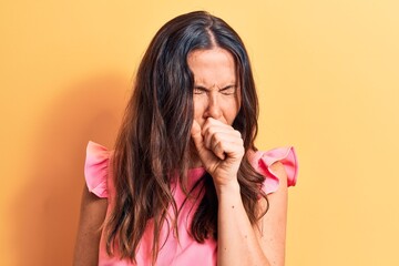 Young beautiful brunette woman wearing pink casual t-shirt standing over yellow background feeling unwell and coughing as symptom for cold or bronchitis. Health care concept.