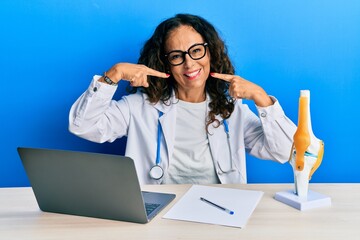 Beautiful middle age woman doctor at orthopedic clinic smiling cheerful showing and pointing with fingers teeth and mouth. dental health concept.
