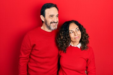 Middle age couple of hispanic woman and man hugging and standing together smiling looking to the side and staring away thinking.