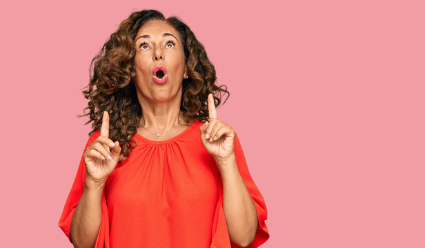 Middle age hispanic woman wearing casual clothes amazed and surprised looking up and pointing with fingers and raised arms.