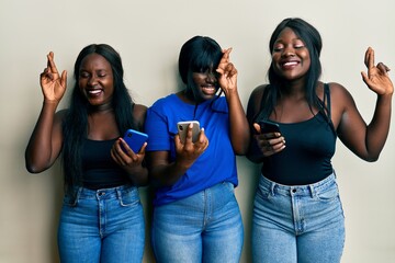 Three young african american friends using smartphone gesturing finger crossed smiling with hope and eyes closed. luck and superstitious concept.