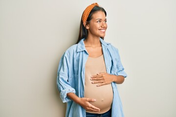 Beautiful hispanic woman expecting a baby, touching pregnant belly looking away to side with smile...