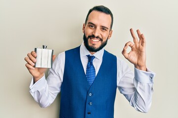 Young man with beard drinking whiskey from flask doing ok sign with fingers, smiling friendly gesturing excellent symbol