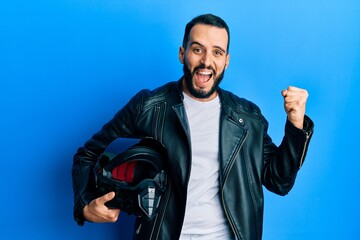 Young man with beard holding motorcycle helmet screaming proud, celebrating victory and success...