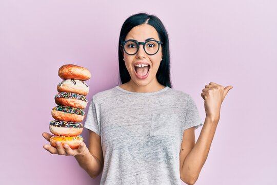 Beautiful young woman holding pile of tasty colorful doughnuts pointing thumb up to the side smiling happy with open mouth