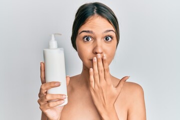 Obraz na płótnie Canvas Young brunette woman holding cosmetic moisturizer facial cream covering mouth with hand, shocked and afraid for mistake. surprised expression