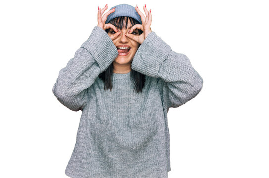 Young hispanic woman wearing cute wool cap doing ok gesture like binoculars sticking tongue out, eyes looking through fingers. crazy expression.
