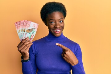 Young african american girl holding 100 new zealand dollars banknote smiling happy pointing with hand and finger