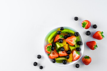 Sliced fresh ripe fruit (strawberries, kiwis, citrus fruits, blueberries) on a white plate. Whole large ripe strawberries and blueberries around the plate. White background.