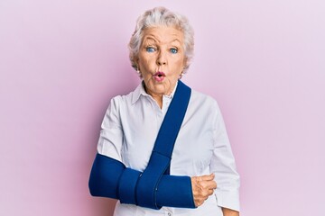 Fototapeta na wymiar Senior grey-haired woman wearing arm on sling scared and amazed with open mouth for surprise, disbelief face
