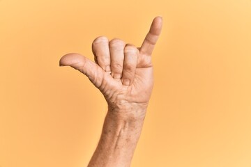 Senior caucasian hand over yellow isolated background gesturing hawaiian shaka greeting gesture, telephone and communication symbol