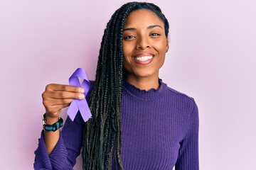 African american woman holding purple ribbon awareness looking positive and happy standing and...