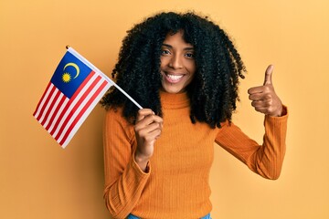 African american woman with afro hair holding malaysia flag smiling happy and positive, thumb up doing excellent and approval sign