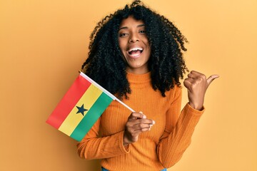 African american woman with afro hair holding ghana flag pointing thumb up to the side smiling happy with open mouth - obrazy, fototapety, plakaty