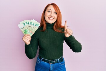 Beautiful redhead woman holding 50 hong kong dollars banknotes smiling with an idea or question pointing finger with happy face, number one