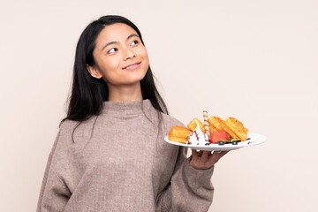 Teenager Asian girl holding waffles isolated on beige background looking up while smiling