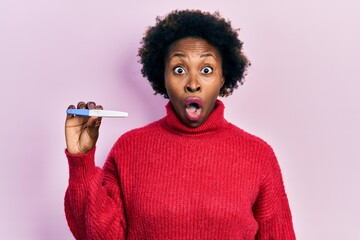 Young african american woman holding thermometer scared and amazed with open mouth for surprise,...