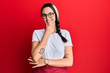 Young hispanic woman wearing professional waitress apron looking confident at the camera smiling with crossed arms and hand raised on chin. thinking positive.