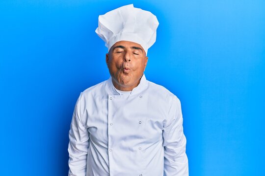 Mature middle east man wearing professional cook uniform and hat making fish face with lips, crazy and comical gesture. funny expression.