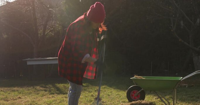 Gardener woman rake leaves dry grass garden lawn meadow spring, Cleaning with rake tool dry grass in the spring, after melting snow. Woman wearing red plaid shirt working in the garden .
